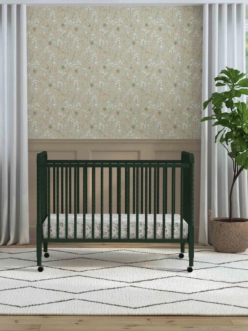 A dark green crib in a nursery with light walls and curtains, a gray armchair, a patterned rug, and a potted plant by the window.