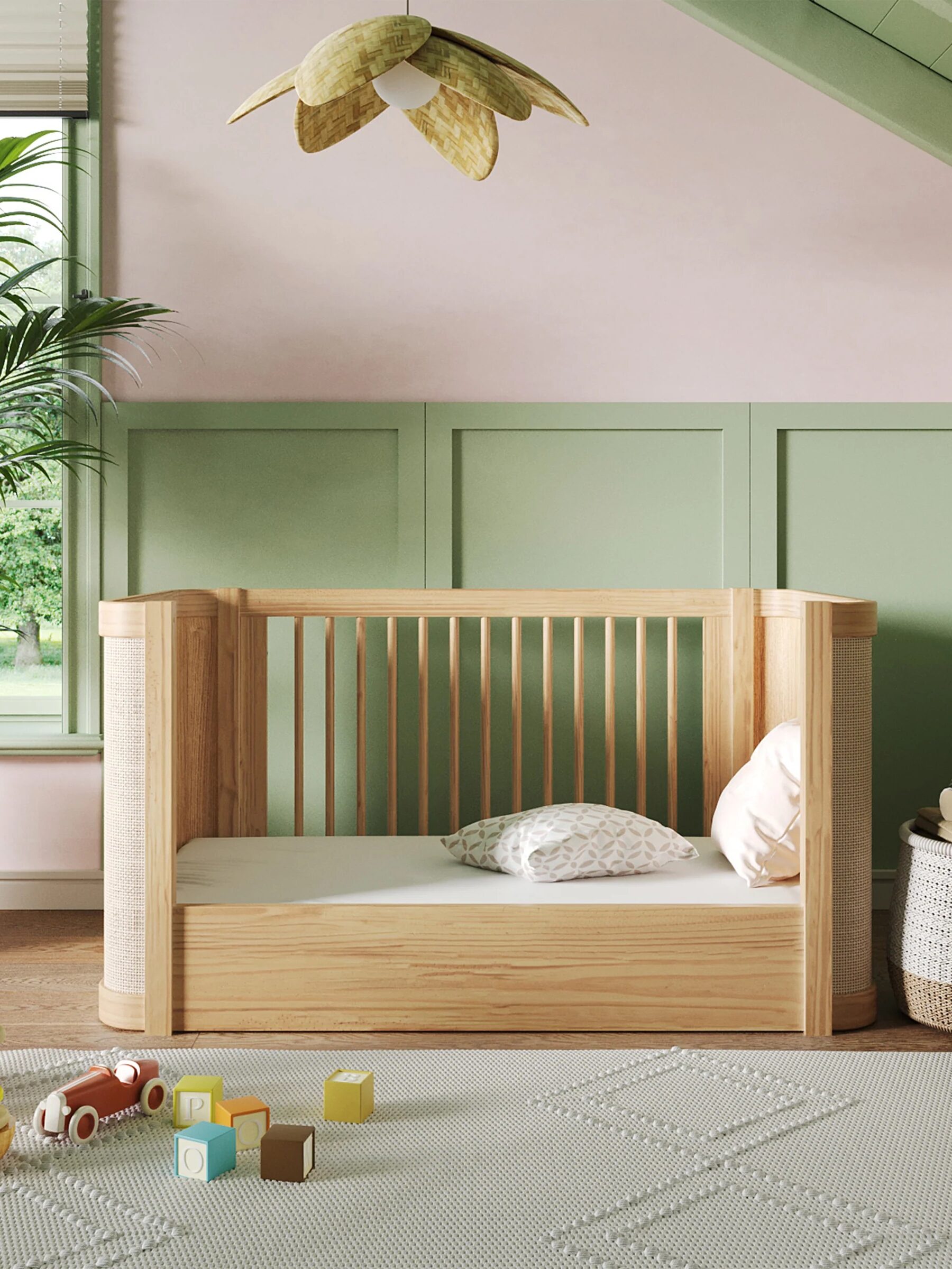 Nursery room with a wooden crib, pastel walls, a rug, and toys on the floor. A plush toy and a basket are in the corner. A ceiling lamp shaped like leaves hangs above.