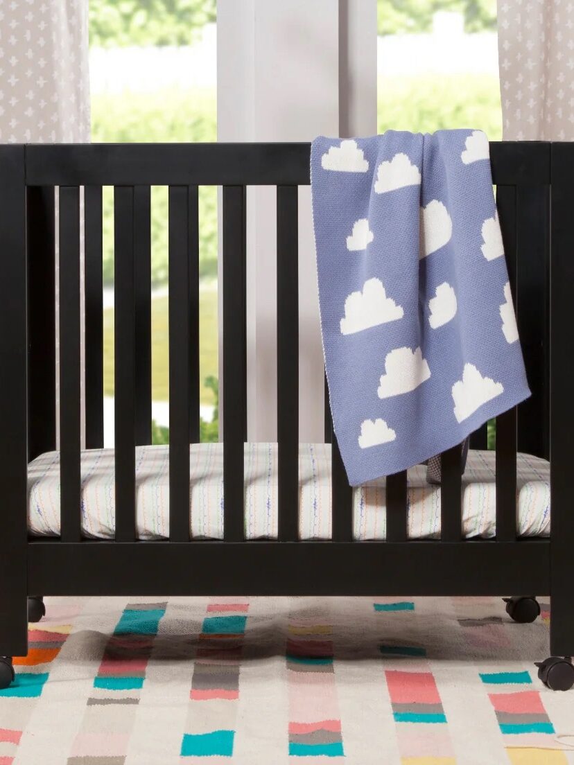 A black crib with a blue cloud-patterned blanket draped over the side. A colorful rug is on the floor, and a shelf with toys and books is in the background.