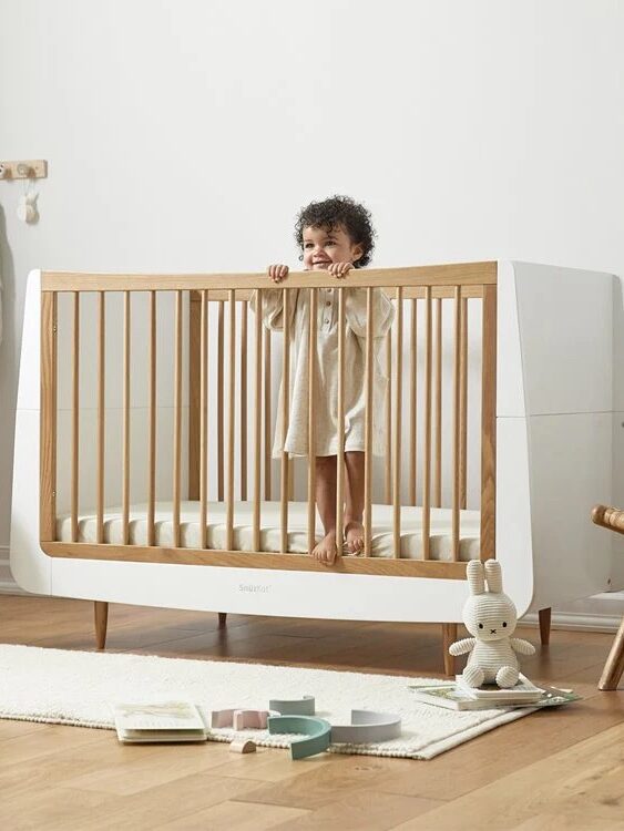 A toddler stands in a wooden crib in a nursery, surrounded by toys and a decorative basket.