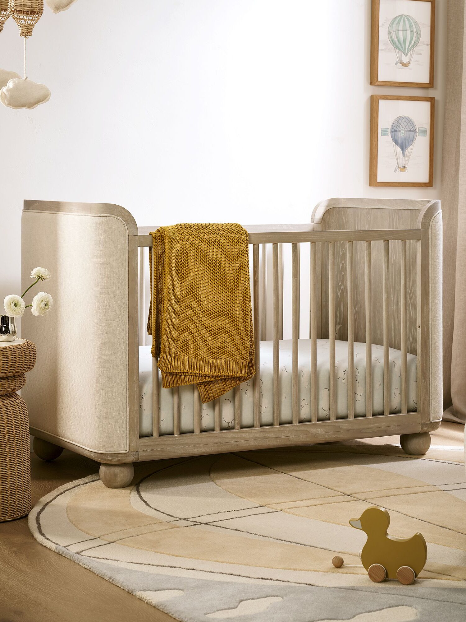 A cozy nursery with a wooden crib, a mustard blanket draped over it, soft decorations, a patterned rug, and a wicker side table with flowers.