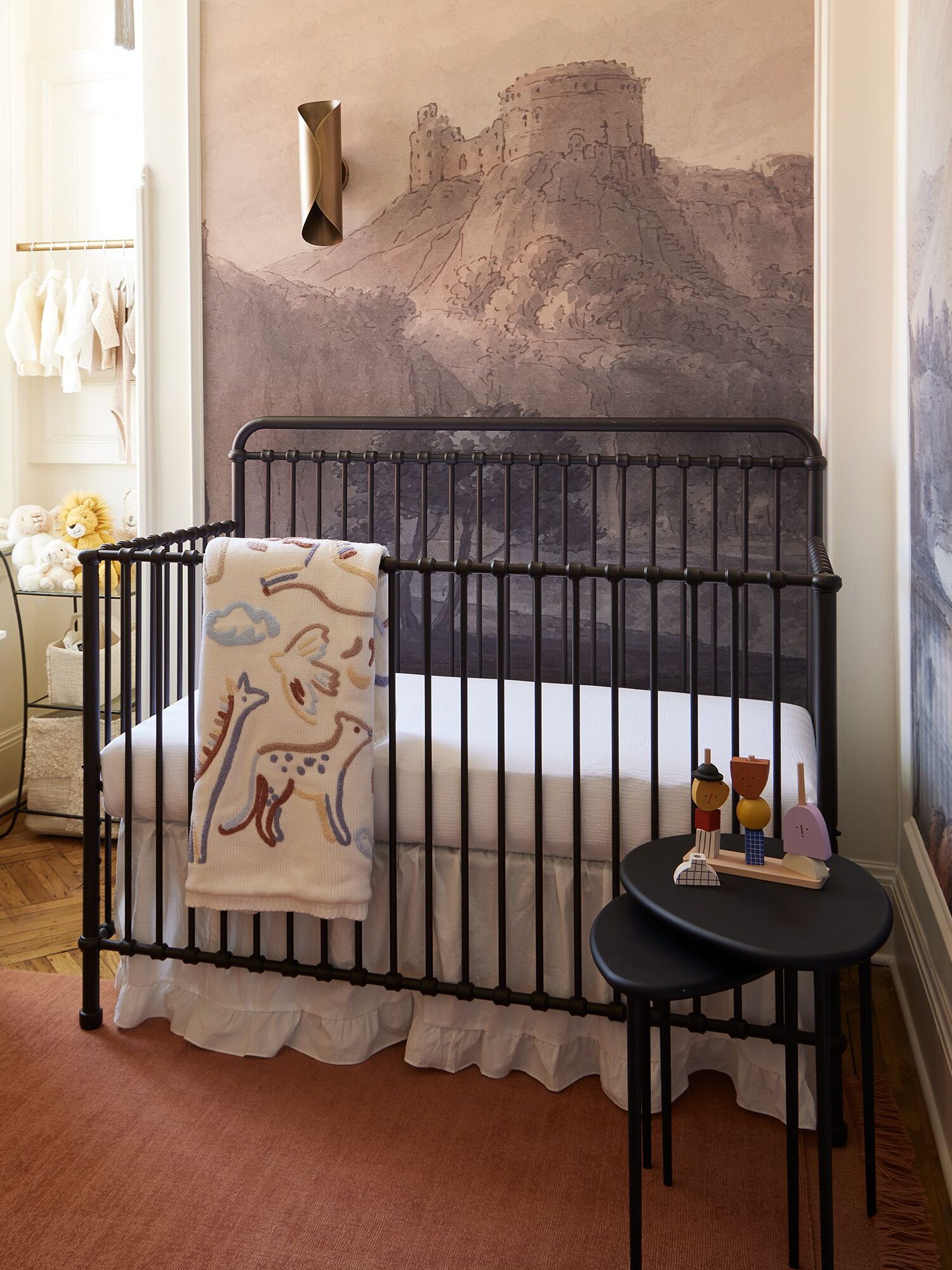 A nursery features a black crib, a tall dresser with a plant, a small round table with decorative items, and a mural of a castle on the wall. A blanket with a playful design hangs on the crib.