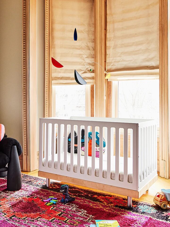 A nursery with a white crib, colorful mobile, and toys on a vibrant rug. A soft chair with a striped heart pillow is nearby, and large windows with beige blinds provide natural light.