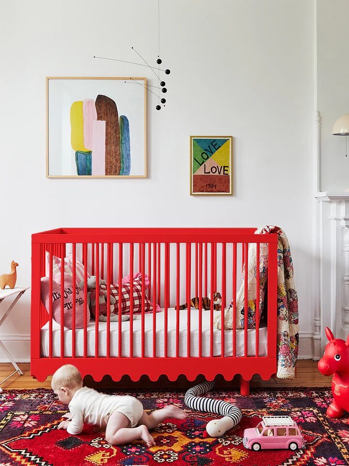 Baby crawling on a colorful rug near a red crib, with toys scattered around. Walls decorated with framed art.