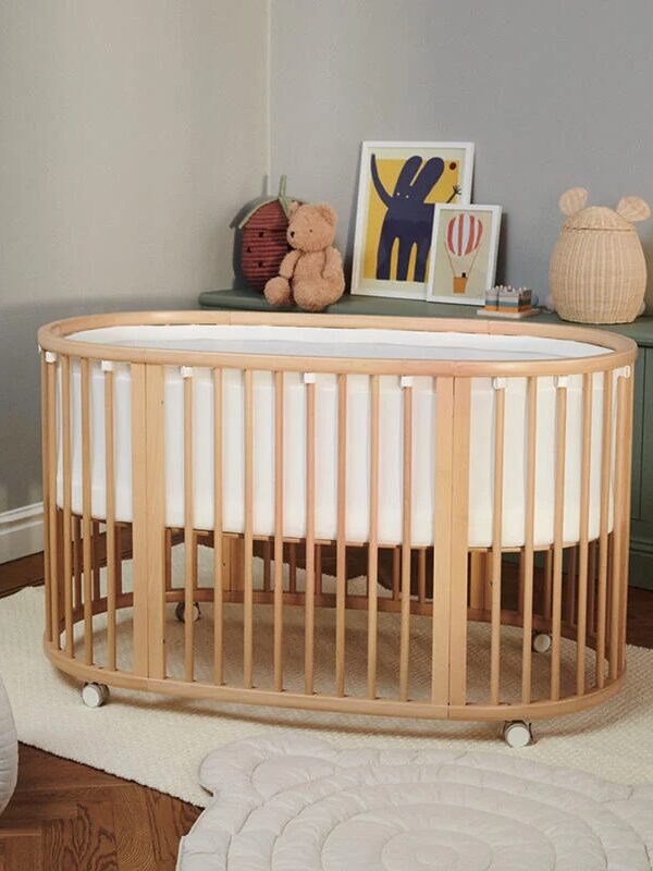 A wooden oval crib with a white mattress is in a nursery. A stuffed teddy bear and art prints are on a green dresser. A plush elephant toy is on the floor near a beige play mat.