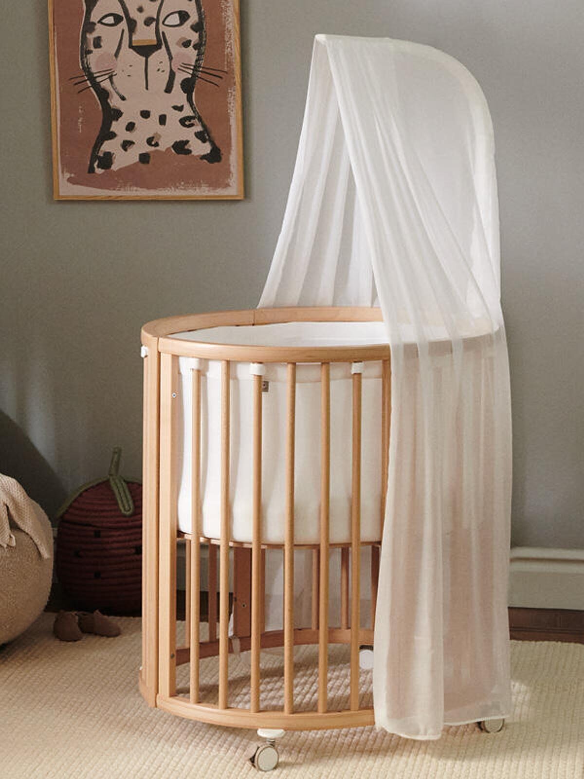 A wooden baby crib with a white canopy in a nursery, next to a plush chair with stuffed toys. A framed animal illustration hangs on the wall.