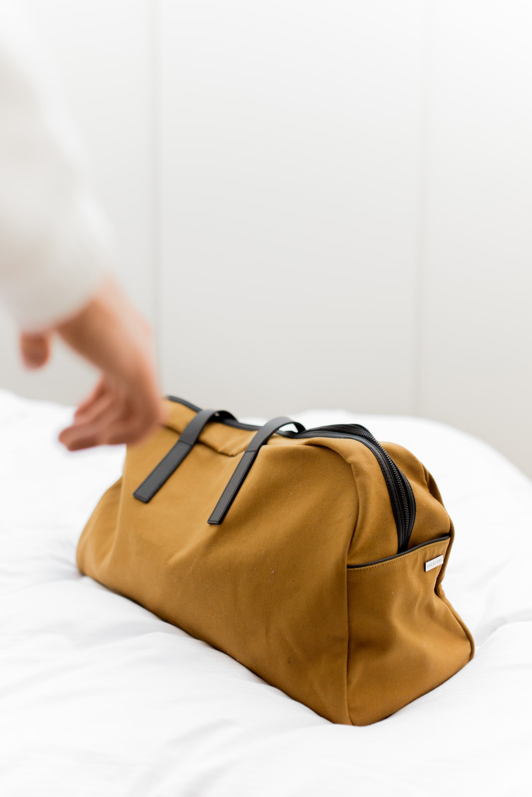 A hand reaches for a brown duffel bag with black handles resting on a white surface.