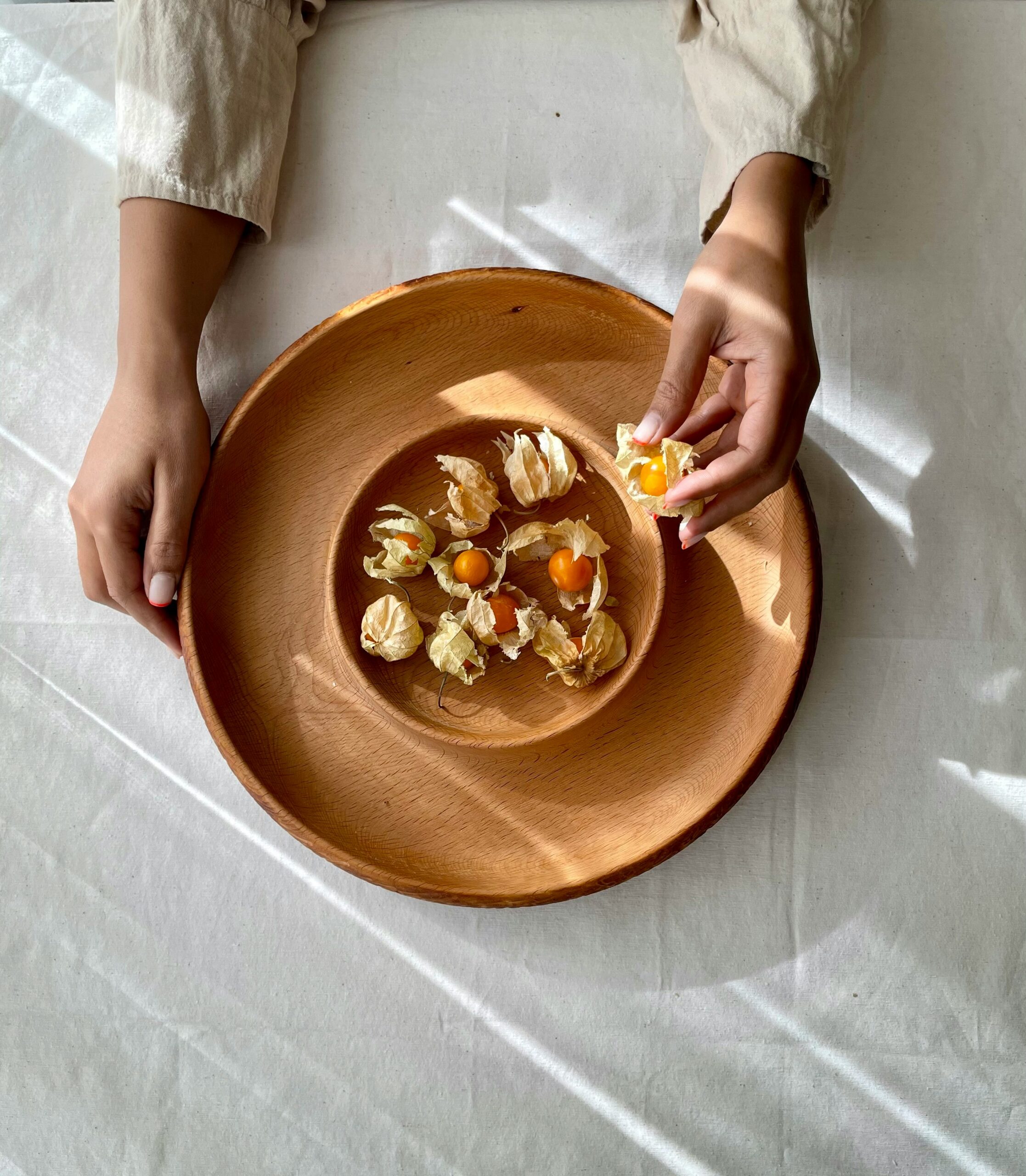 Person peeling husk cherries on a round wooden plate, with sunlight creating shadows on a white tablecloth.