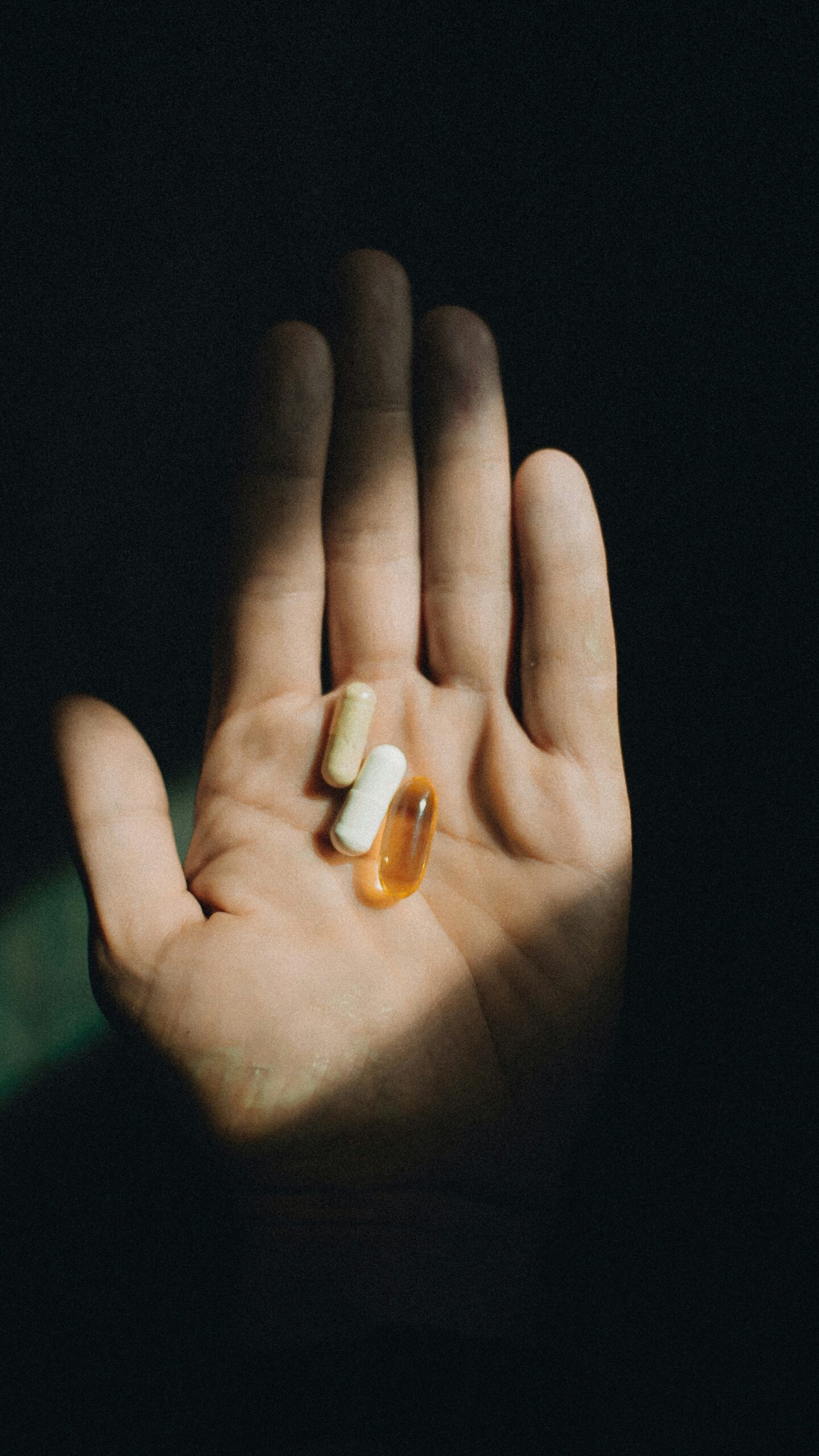 An open hand holding three pills of different shapes and colors against a dark background.