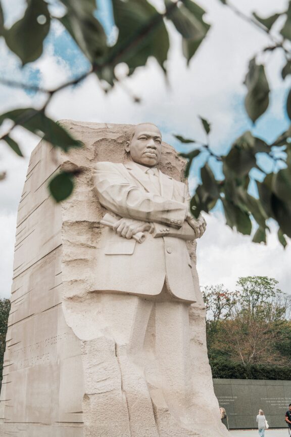 A large stone monument of a man in a suit, with arms crossed, stands against a background of trees and sky. Leafy branches partially frame the top of the image.