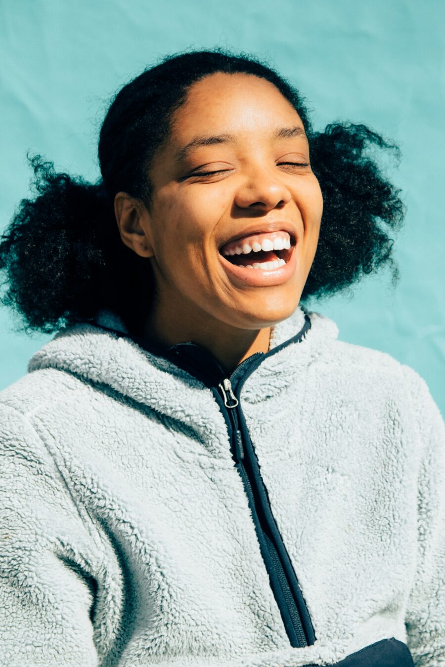 Person with curly hair wearing a light fleece jacket smiles broadly against a turquoise background.