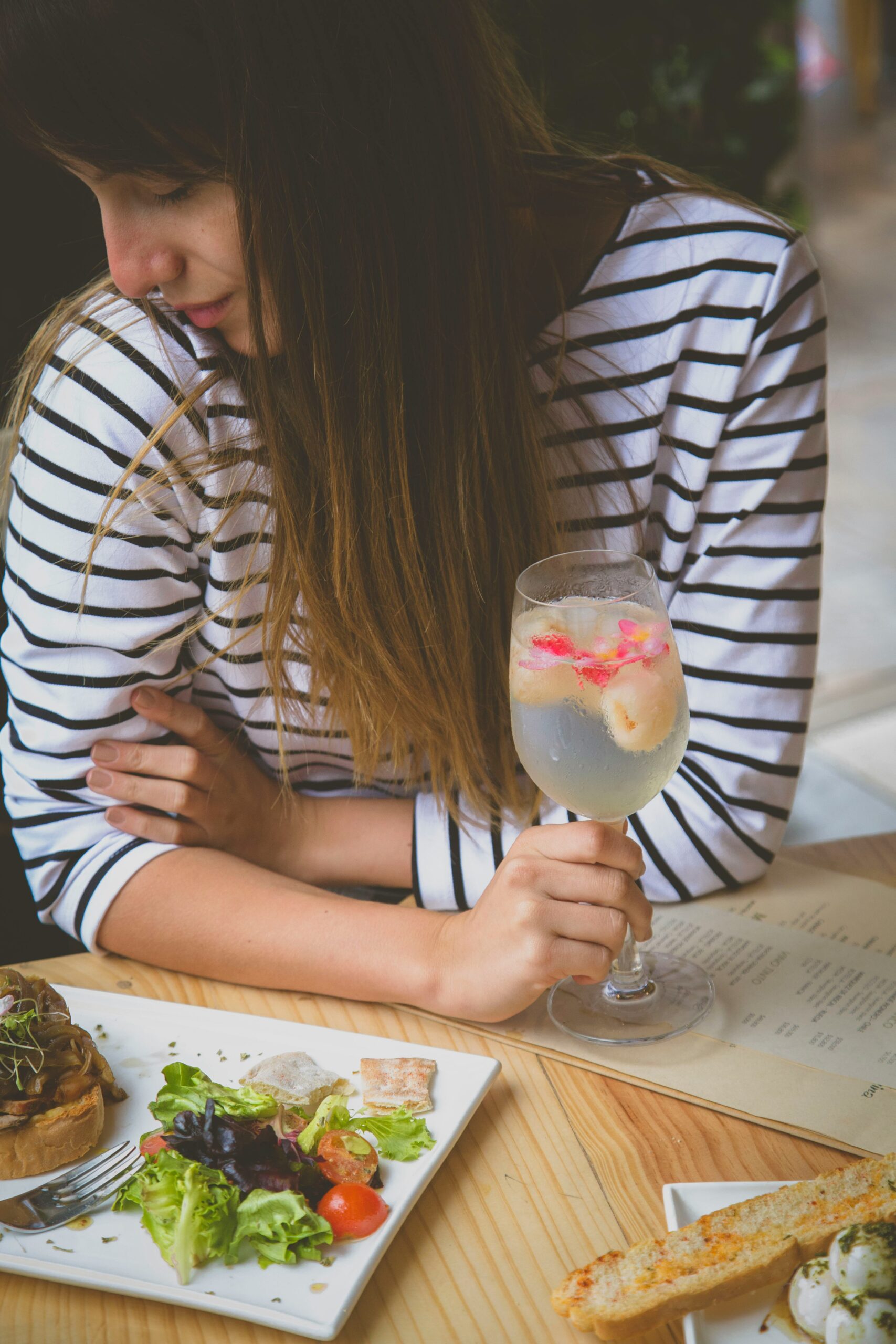 Person in a striped shirt sitting at a wooden table, holding a glass with floral garnish. Plates of salad, tart, and breadsticks are on the table.