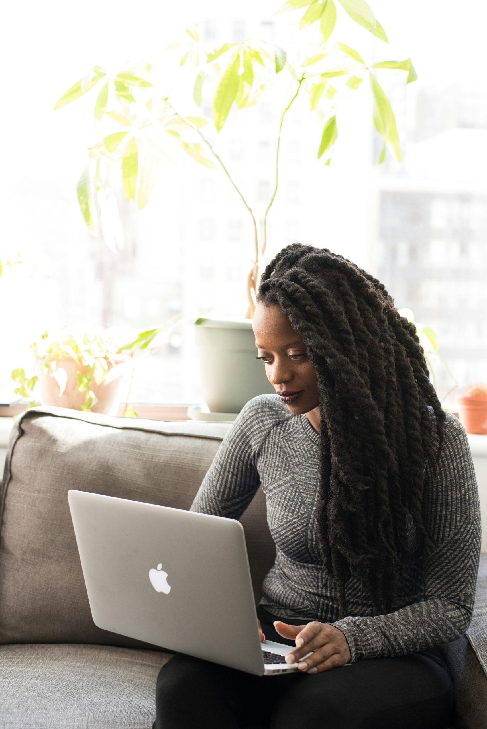 Person with long hair works on a laptop while seated on a couch, in a brightly lit room with plants in the background.