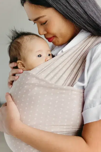 A woman gently holds a baby in a beige baby wrap, looking down with a soft smile.