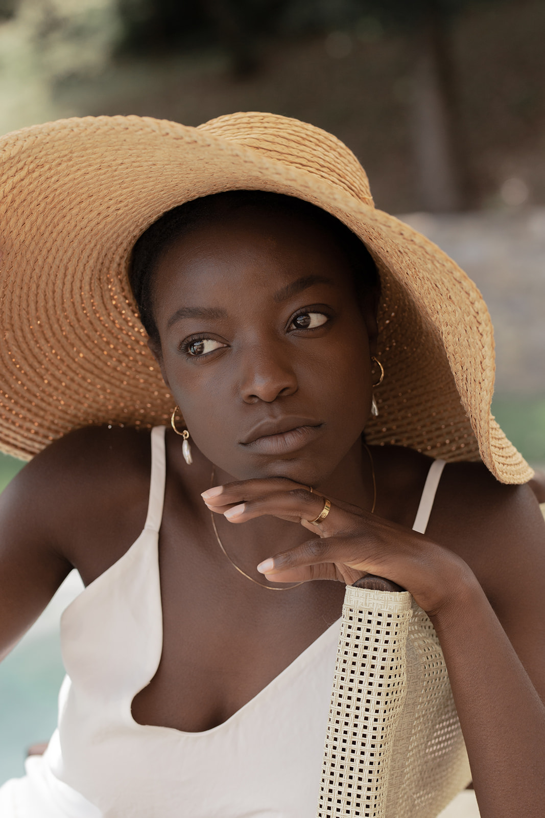 A woman in a white dress and large straw hat gazes thoughtfully to the side, her chin resting on her hand. She is outdoors in a softly lit setting.