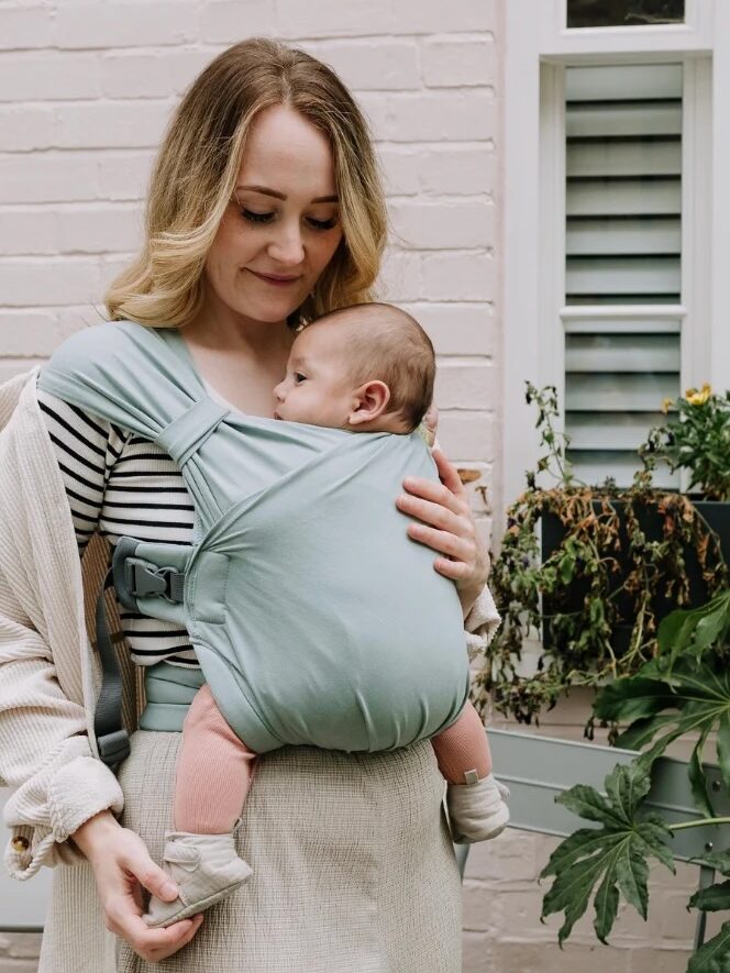 A woman with long hair carries a baby in a blue baby carrier. She stands in front of a brick wall and a window with plants nearby.