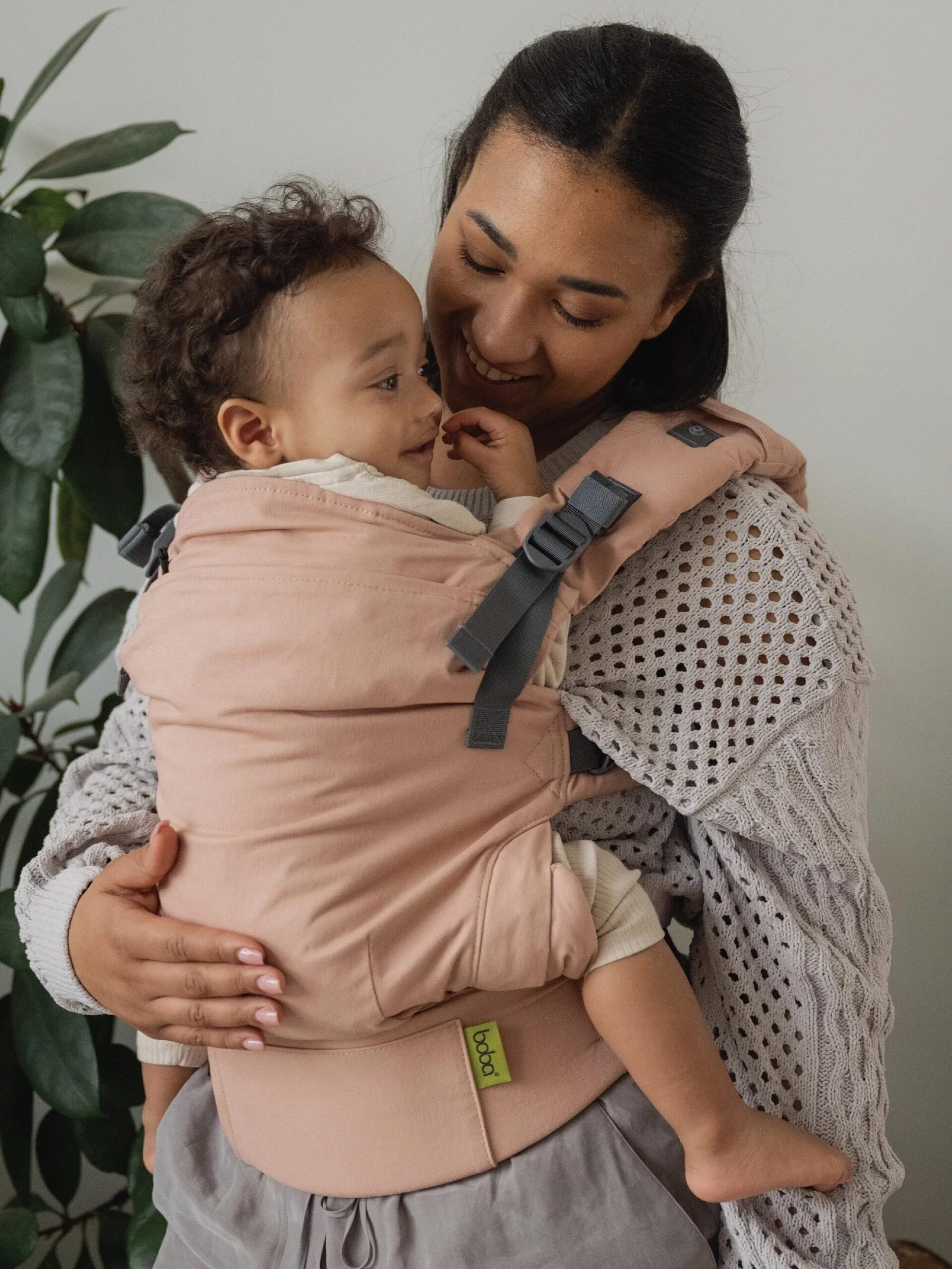 A woman holds a smiling baby in a pink baby carrier, standing in front of a green plant.