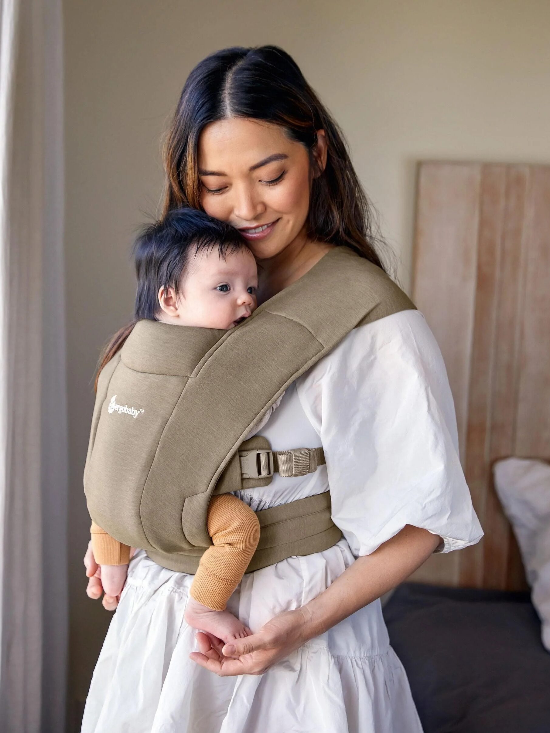 A woman stands indoors, smiling down at a baby secured in a brown baby carrier on her front. The woman is wearing a white dress.