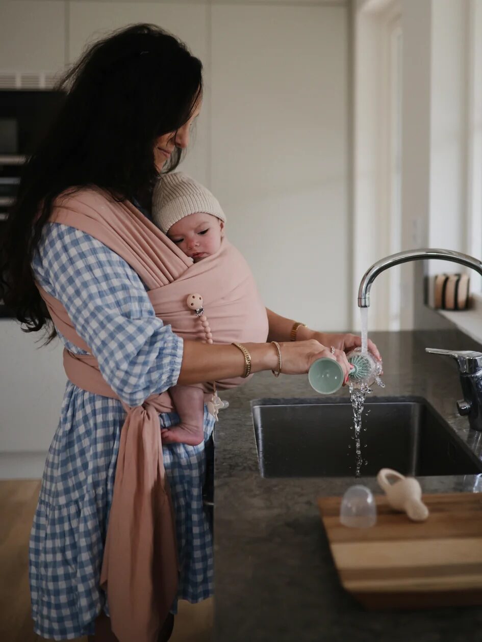 A woman holds a baby in a wrap while standing in a kitchen next to a sink. She is wearing a blue and white checkered dress.