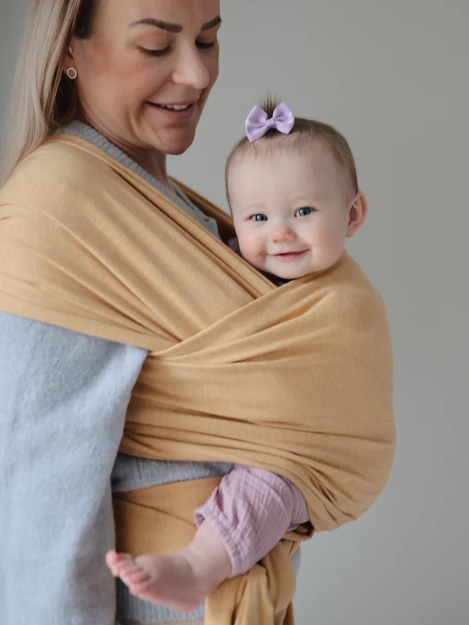 A woman carries a smiling baby wrapped in a yellow baby sling.