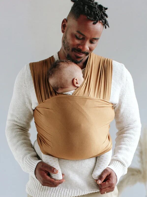 A man stands holding a baby in a brown baby wrap. He is looking down at the baby with a gentle expression.