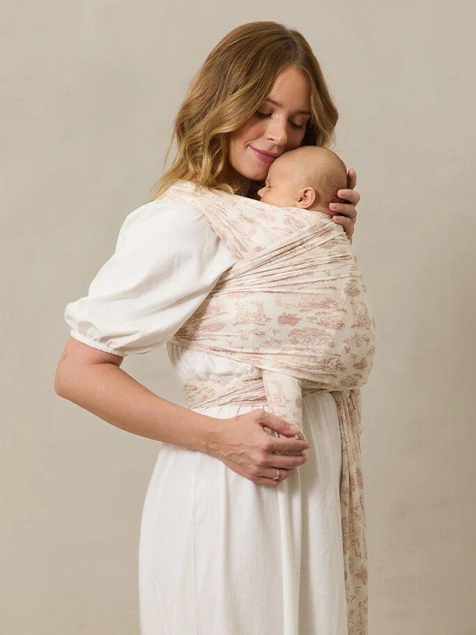 A woman in a white dress holds a baby wrapped in a patterned baby carrier against her chest.