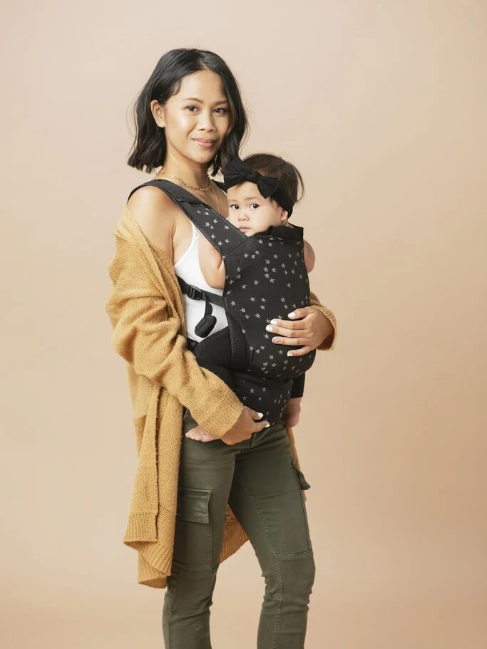 Woman in a tan sweater and green pants holding a baby in a black star-patterned carrier against a beige background.