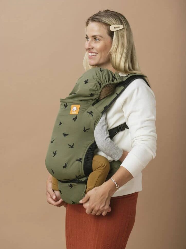 A woman stands smiling, carrying a baby in a green baby carrier with black patterns. She is wearing a white sweater and red skirt against a plain beige background.