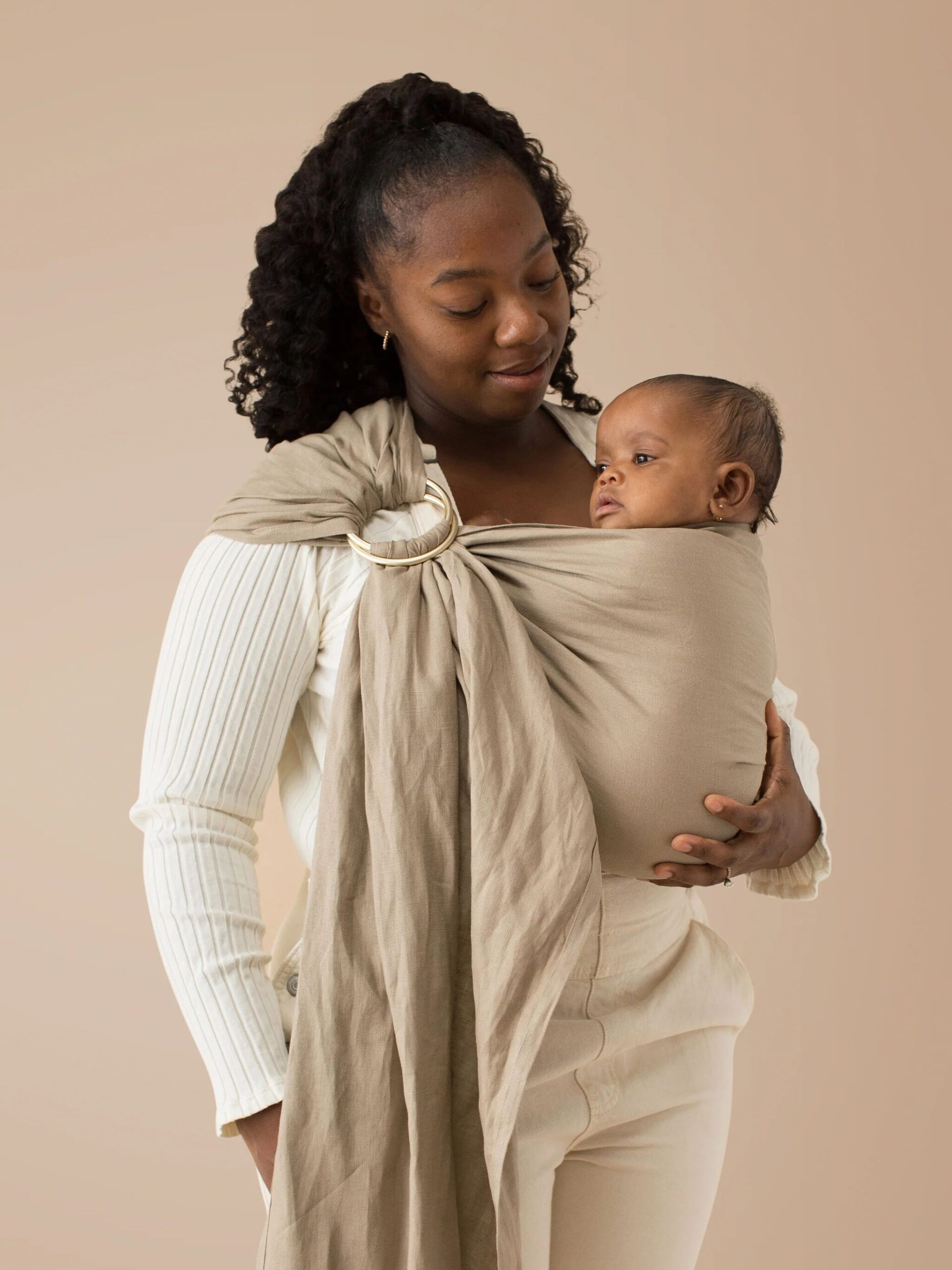 A woman in a white shirt holds a baby in a beige sling against a neutral background.