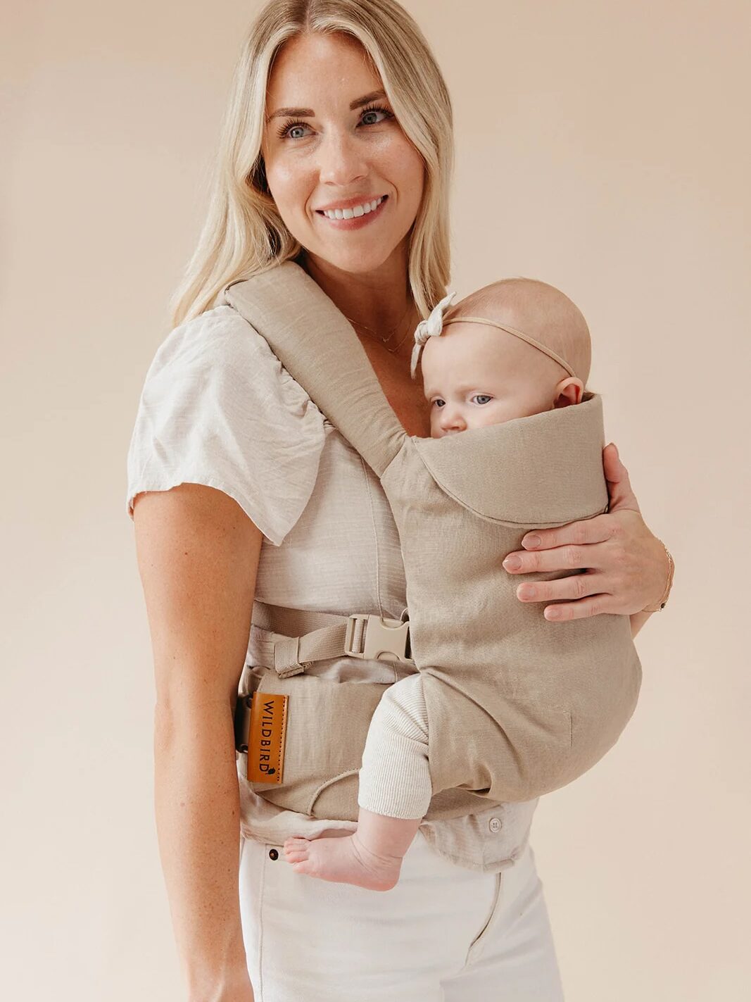 A woman wearing a beige top and white pants holds a baby in a beige baby carrier. The baby has a bow on their head.