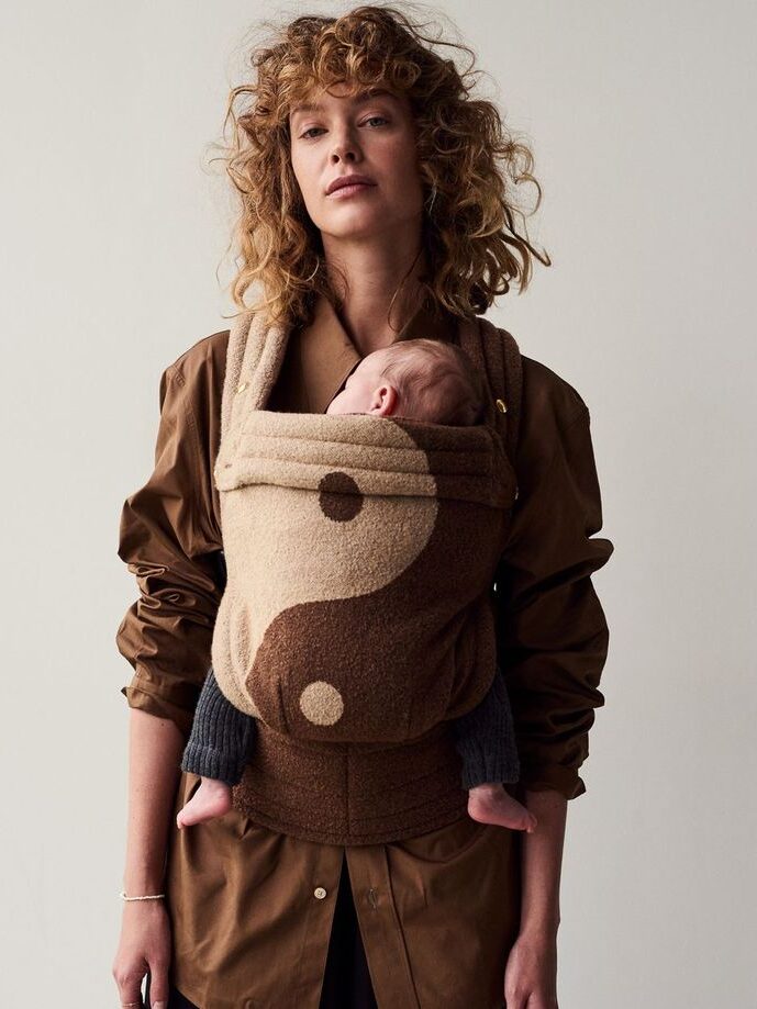 A person stands against a plain background, holding a baby in a brown yin-yang patterned carrier.