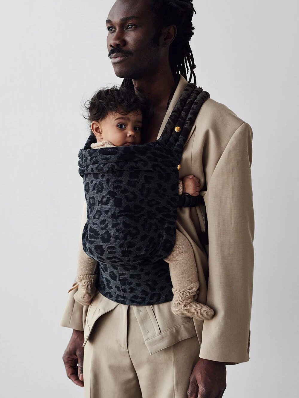 A person in a beige suit carries a baby in a leopard-print carrier, standing against a plain background.