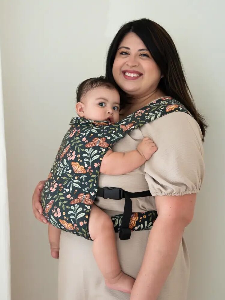 A woman wearing a beige dress carries a baby in a floral-patterned baby carrier.