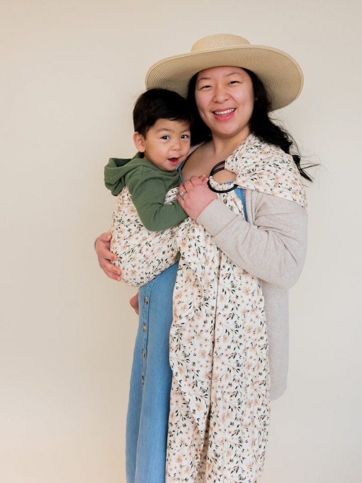 A woman in a hat carries a smiling child in a floral wrap.