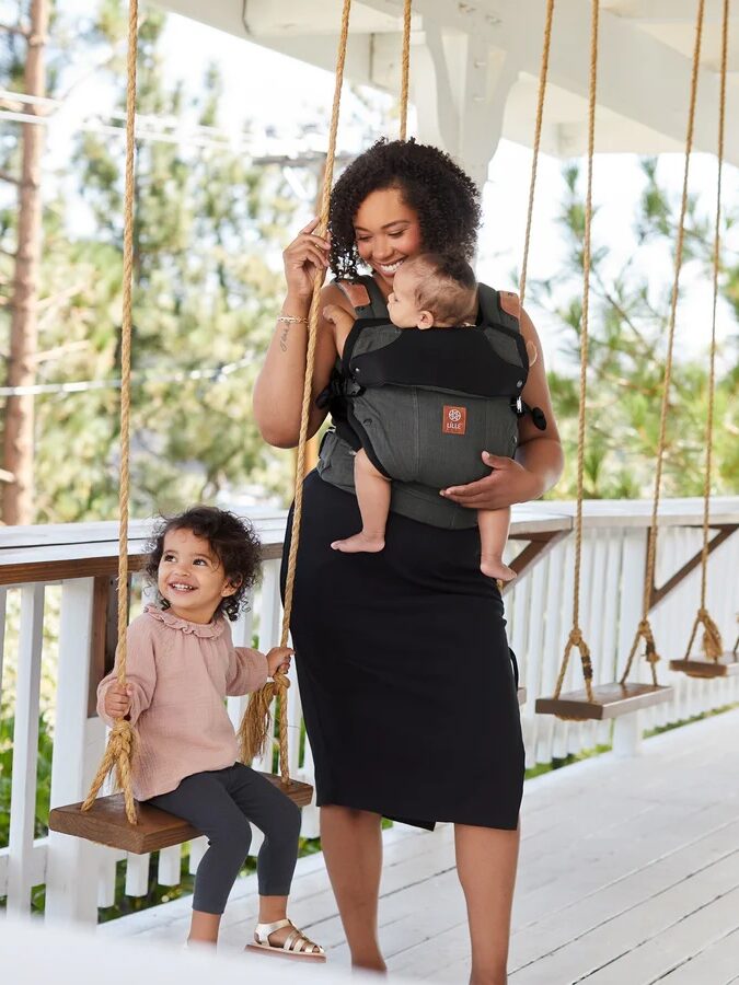 A woman stands on a porch, holding a baby in a carrier. A young child sits on a swing nearby, smiling. The background features trees and a wooden railing.