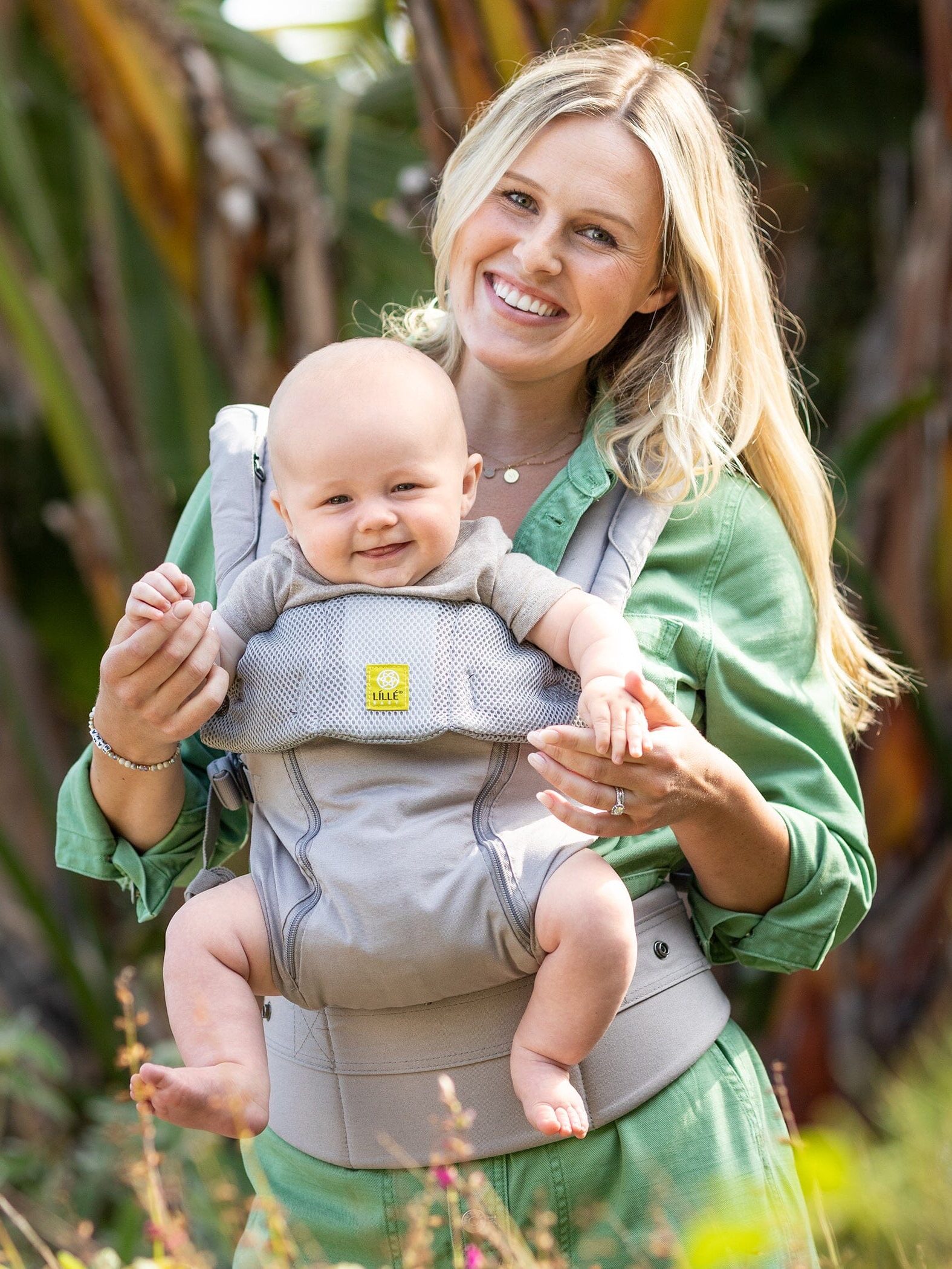 Woman in a green dress carrying a smiling baby in a gray baby carrier outdoors near plants.