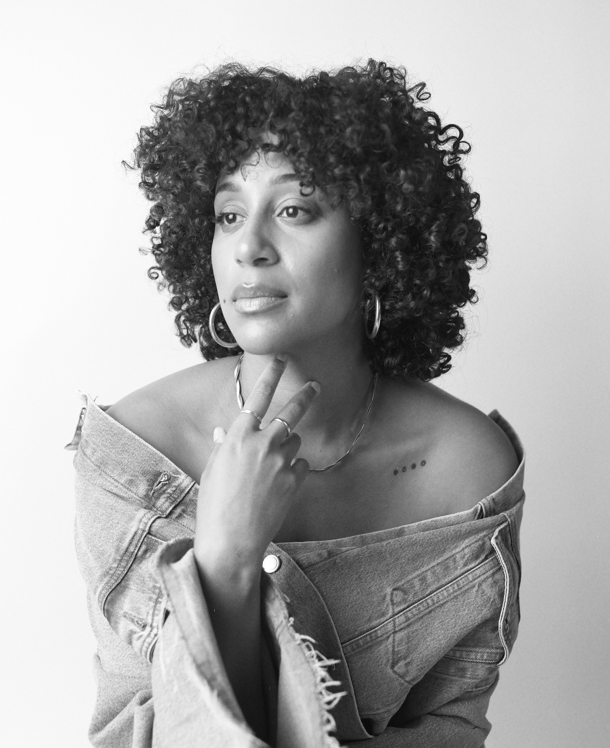 Black and white portrait of a person with curly hair, wearing an off-shoulder denim jacket, looking to the side with a thoughtful expression.