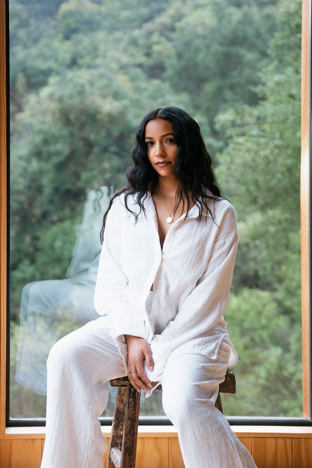 A person in a white outfit sits on a wooden stool in front of a large window, with green foliage visible outside.