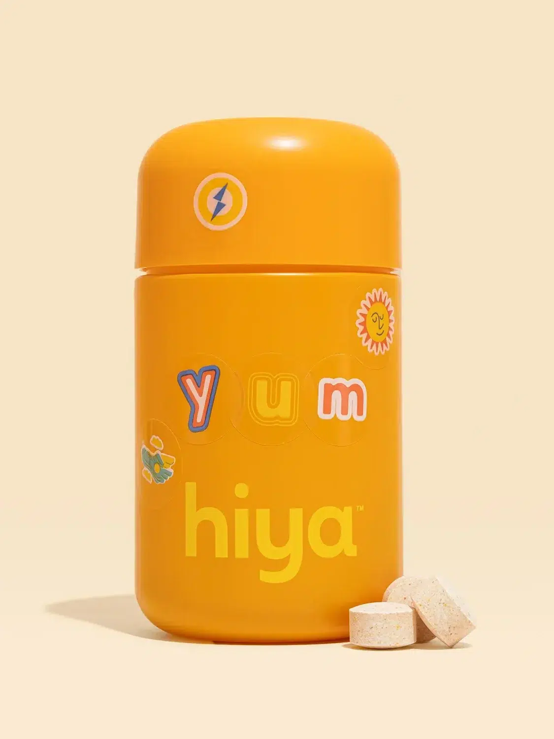 Bright orange bottle with the text "hiya" and colorful stickers, next to two round tablets on a beige background.