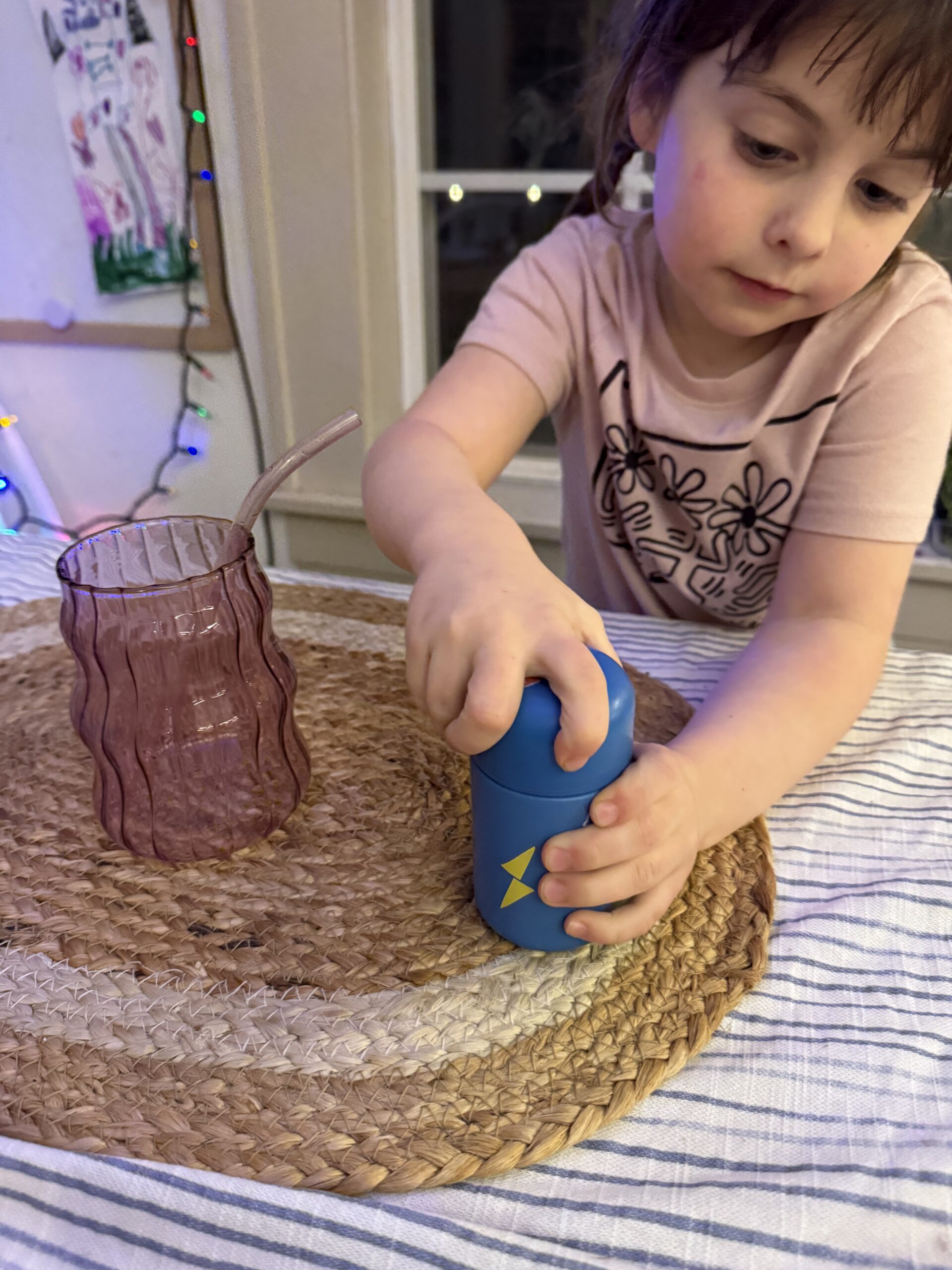 Child opening a blue can next to a purple glass on a woven mat.