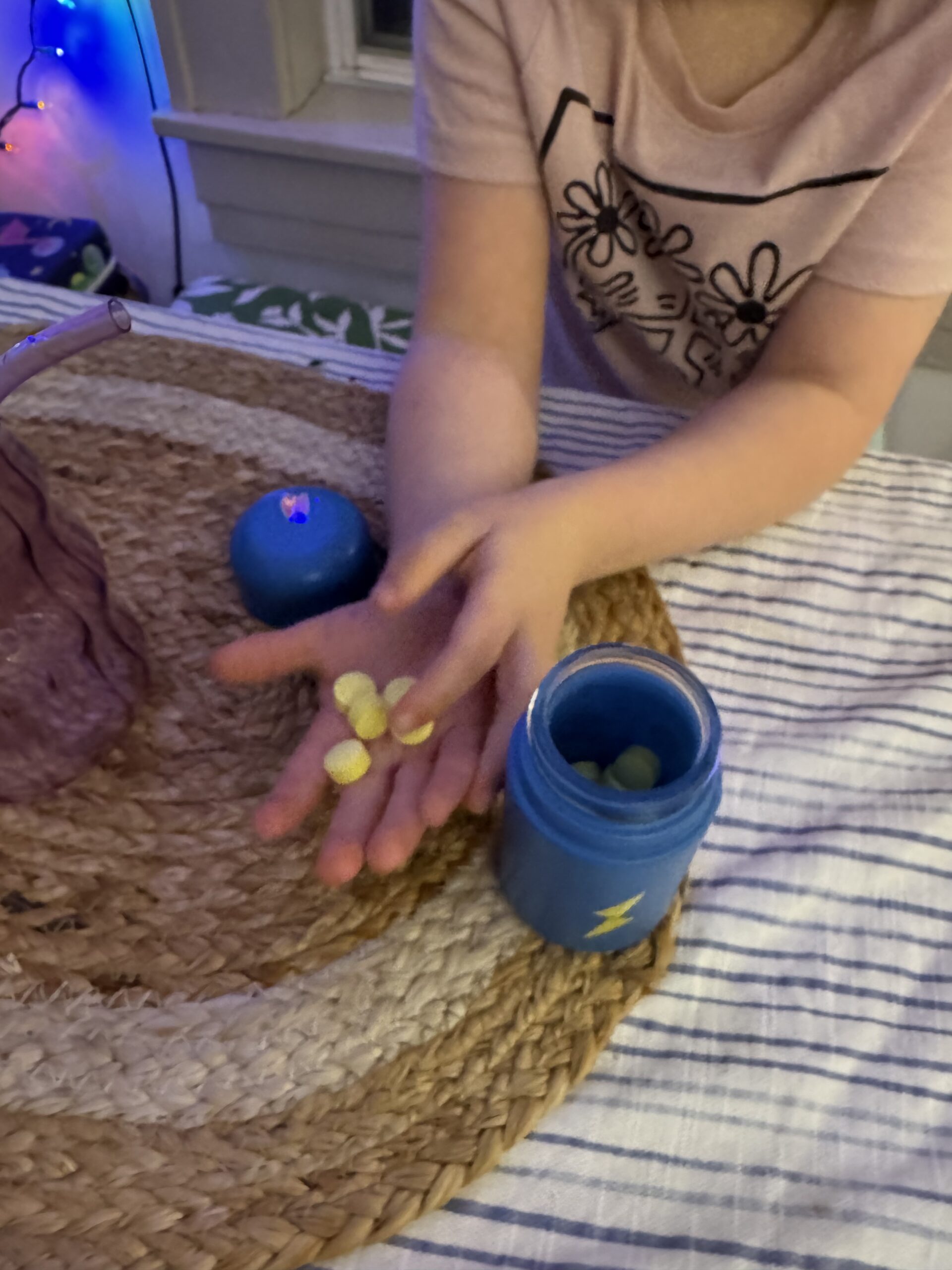 A child holds small round objects in their hand, next to a blue container on a woven mat.