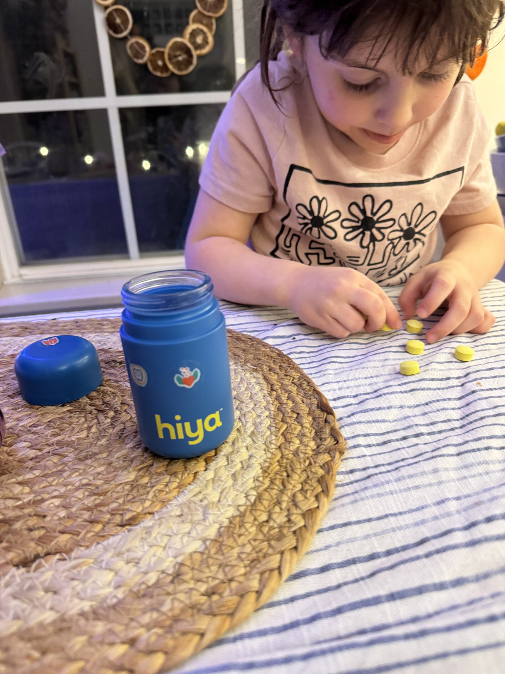 Child playing with yellow tablets next to a blue "hiya" bottle on a table with a striped cloth and woven mat.