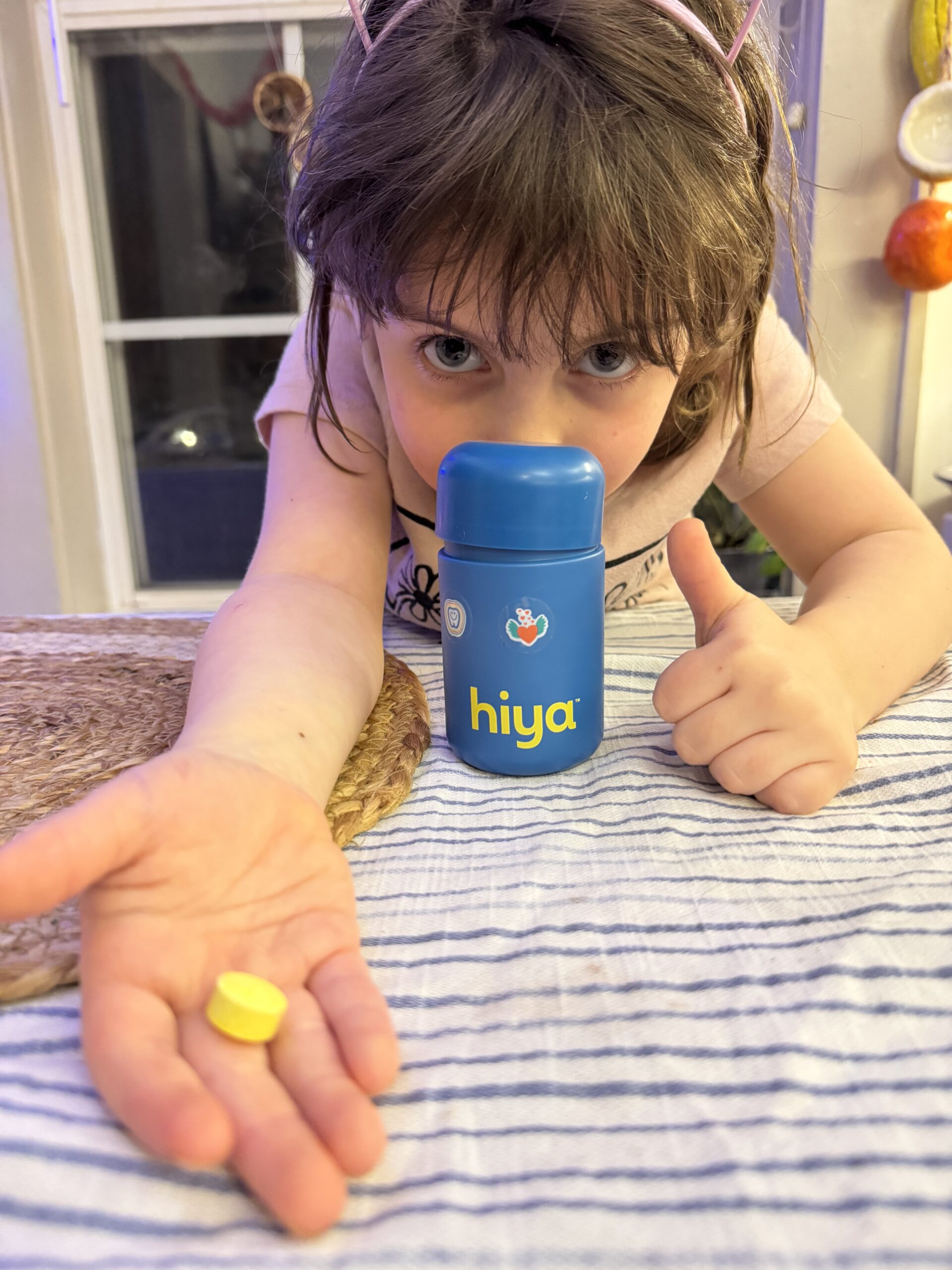 Child wearing cat ear headband giving a thumbs up, holding a yellow tablet, and pointing at a blue "hiya" bottle on a striped tablecloth.