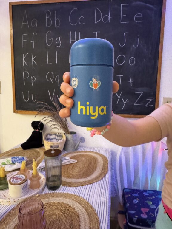 A child holding a blue container labeled "hiya" in a classroom setting with an alphabet chart on a chalkboard in the background.