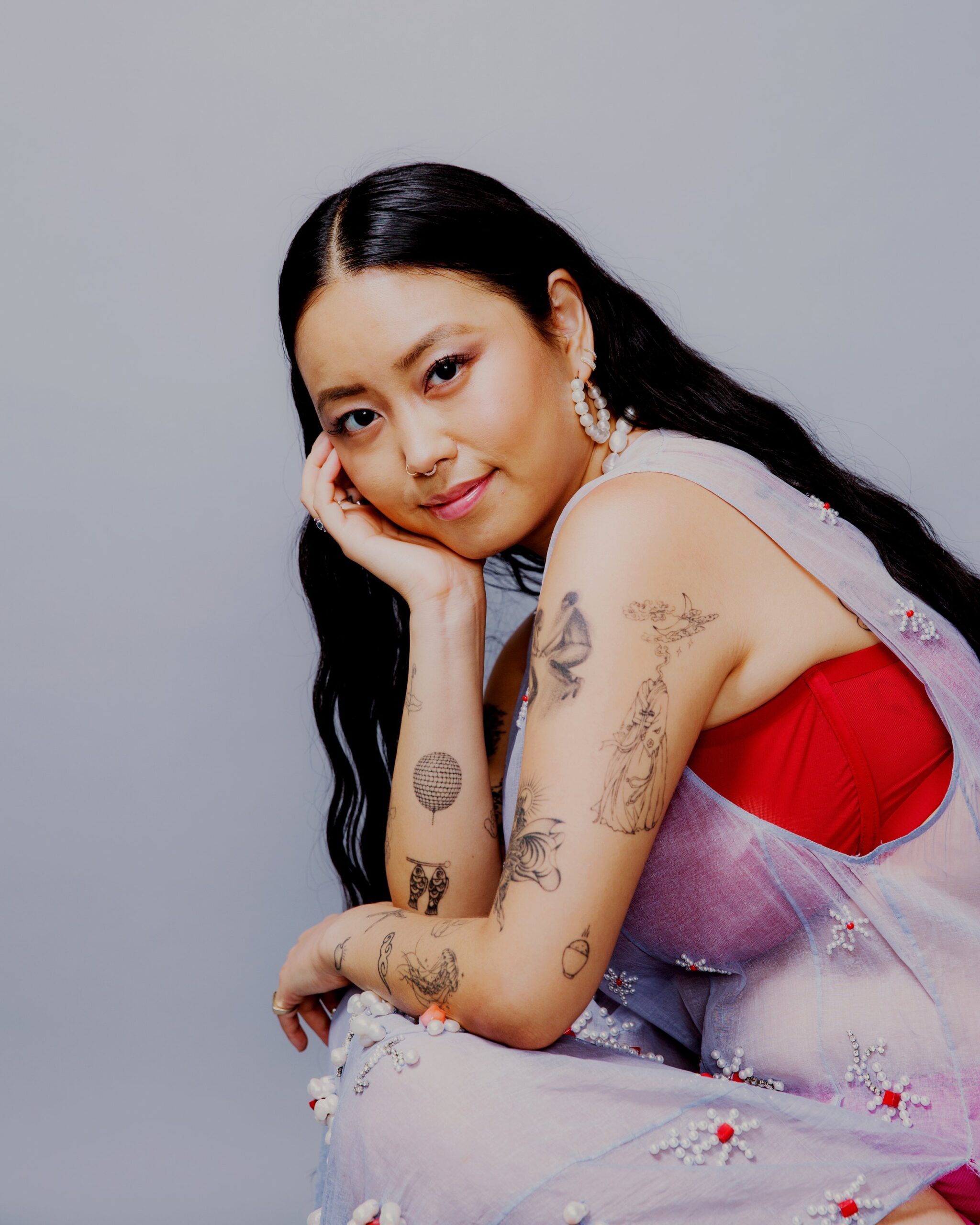 Person with long black hair and various tattoos is seated against a light background, wearing a sheer, embroidered garment over a red top.