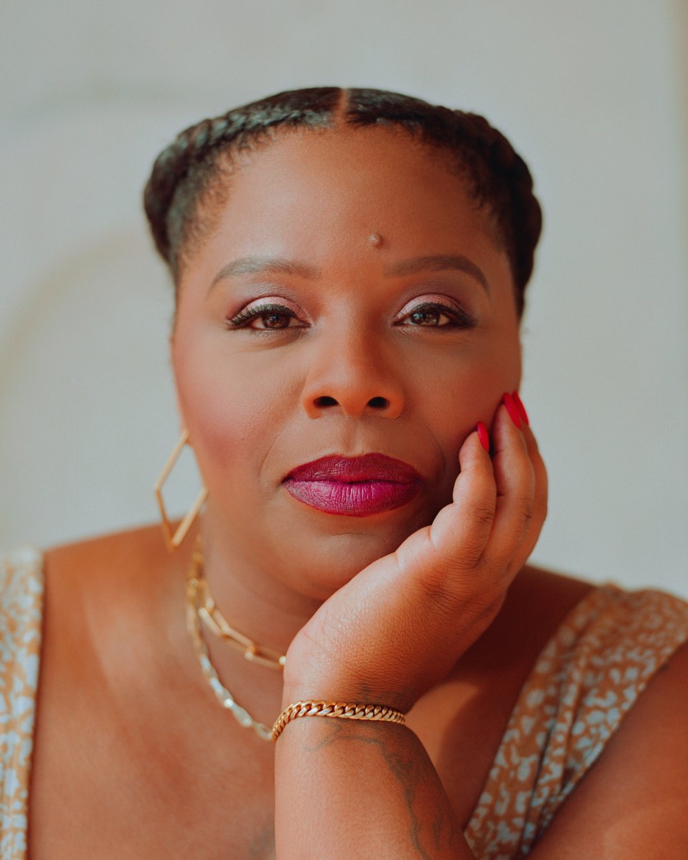 A woman with braided hair rests her chin on her hand, wearing red lipstick and large earrings.