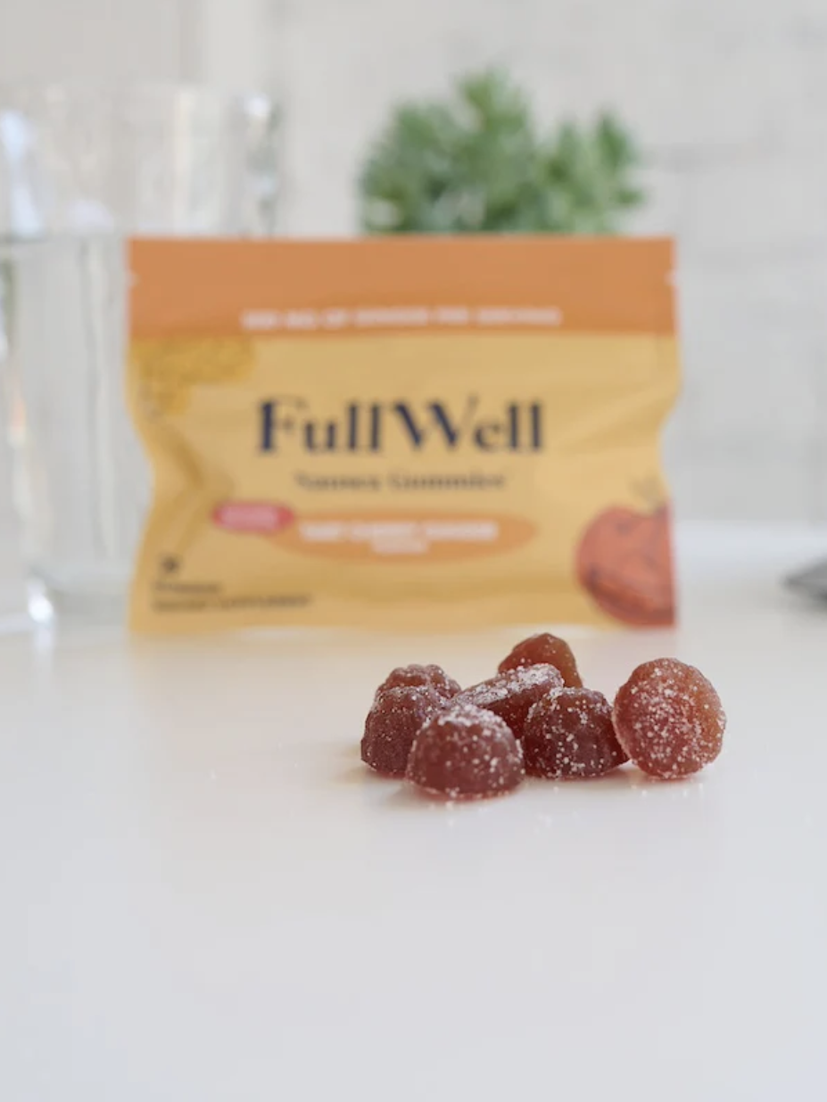 Pile of red gummy vitamins on a white table in front of a FullWell package. A glass and small plant are blurred in the background.