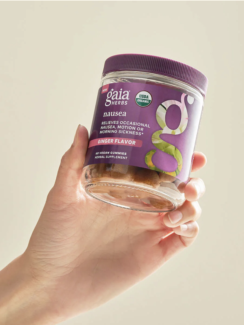 A hand holding a jar of Gaia Herbs Nausea Vegan Gummies, ginger flavor, labeled for relieving nausea, motion, or morning sickness. The jar has a purple lid and label.