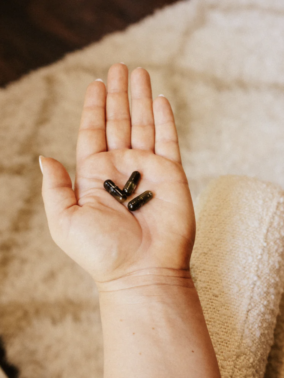 An open hand holding three black capsules, resting over a textured beige surface.