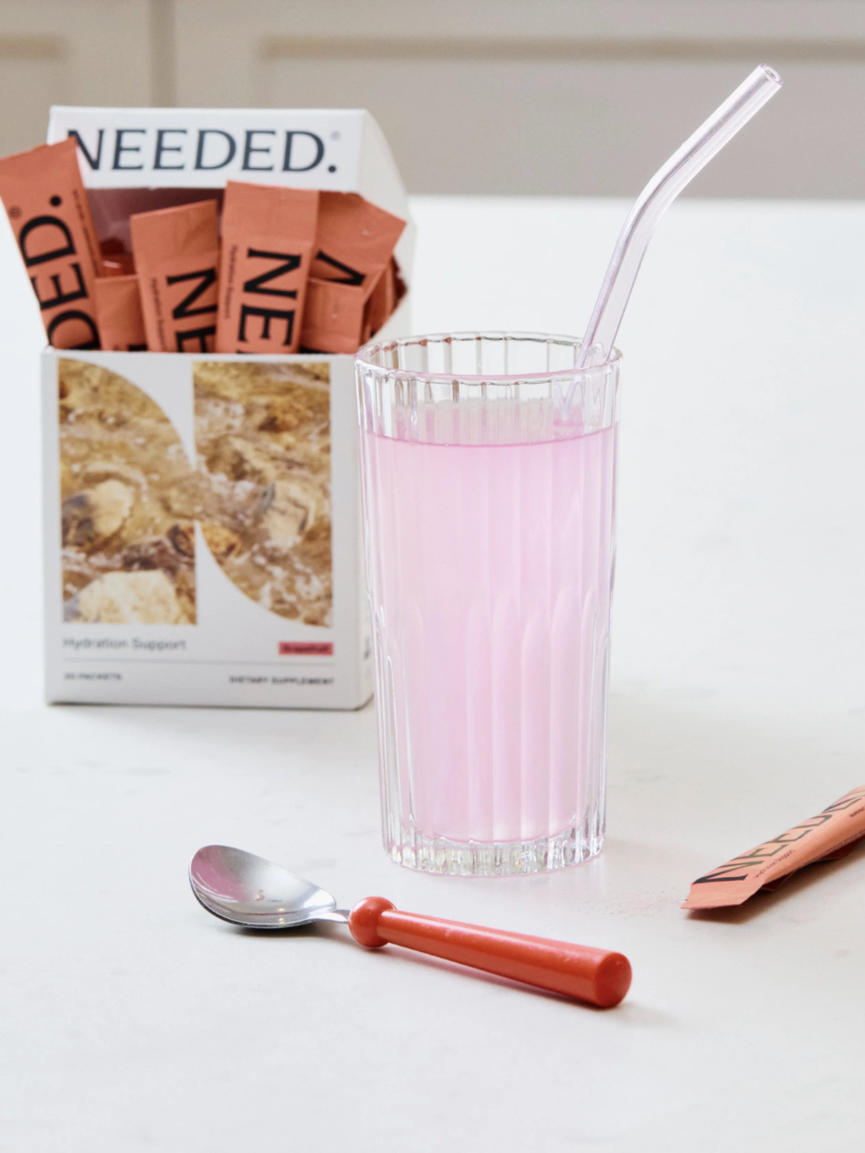 A box of nutritional supplement packets labeled "NEEDED" is on a table next to a glass of pink liquid with a straw and a red-handled spoon.